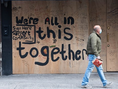 Photo of a man wearing a surgical mask walking past a graffiti which reads: We're all in this together